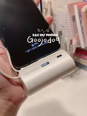 A hand holds a white rectangular power bank connected to a black smartphone via a white cable. The phone displays a battery icon showing 59% charge with the text "1 giờ 1 phút nữa sẽ đầy". Overlayed on the phone and slightly opaque is the text "SẠC DỰ PHÒNG GooJojog". The scene takes place indoors with soft, warm lighting, suggesting a product demonstration or review.