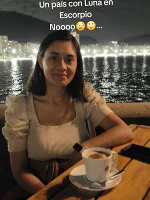 A woman sits at a table, likely outdoors at night. She's in front of a body of water with city lights in the background. She's holding a cup of coffee. Text is overlaid on the top of the image.