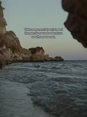 An image showing a tranquil beach scene. In the foreground, dark blue waves gently roll towards the shore. To the left, weathered, brown rocks form the shoreline. Behind these rocks, a cliff face rises. Overhead, the sky transitions from a light, almost creamy tone at the horizon to a darker, muted blue at the top, suggesting either dusk or dawn.