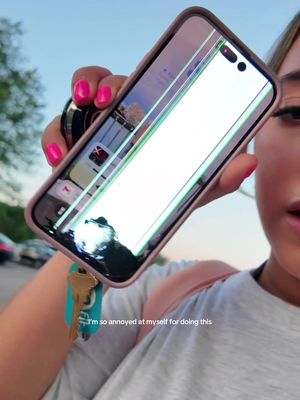 A woman holds a cracked iPhone, the screen displaying a bright white section with green and white lines and a dark broken area. She holds the phone up with pink nails in front of a blurry outdoor background. The video overlays a caption text that is at the bottom.