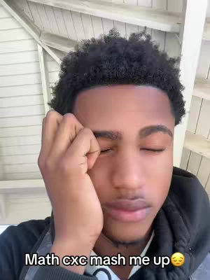 A young man with dark skin and curly black hair is the primary focus. He's wearing a black hooded jacket, and his right hand is gently touching his face, near the closed eyes. The background includes visible elements of a light-colored ceiling and wall.