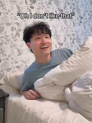 A young man lies in bed, with white bedding and floral wallpaper in the background. The man is smiling and speaking, with text displayed over his head. He is holding the covers up to his chest.
