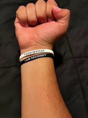 A close-up shot of a clenched fist with two wristbands. The top wristband is white and reads "GOD IS FIRST +" with a cross symbol. The bottom wristband is black and reads "I AM SECOND +" with a cross symbol. The background is a blurred dark surface. The forearm is visible.