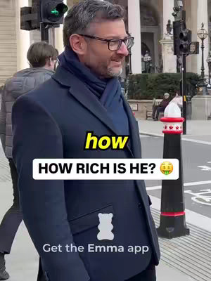 A man in a dark overcoat and glasses stands on a city street, looking to the side. Overlayed text asks, "HOW RICH IS HE?" with a yellow "how" above. A cartoon gummy bear graphic is below the overlay. More in the background are traffic lights, buildings, and a person walking.