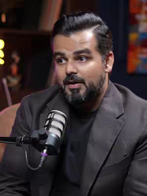 A medium shot shows a man seated in a dimly lit room, facing slightly to the left. He has dark hair neatly styled to the side with a beard and mustache. He wears a dark blazer over a black shirt, and the microphone is positioned in front of him with a background that includes bookshelves and soft lighting.