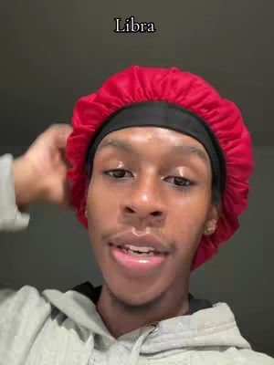 A selfie-style shot features a young Black man. He wears a red durag and a light-colored hoodie. He is looking towards the camera, seemingly talking. The background is a muted grey. Above his head, the word "Libra" is written in white font.