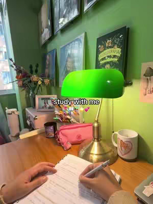 The image shows a study scene with a desk and various items, including a green lamp, a mug, books, and a pink pencil case. Two hands are visible, one holding a pen and the other on a notebook with visible text. The background is dominated by green walls and pictures.