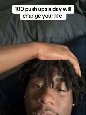 A selfie-style shot featuring a young man; he is positioned in the lower half of the frame, and his head is tilted upwards, gazing towards the camera. Above his head is a white rectangular text box. The background is a dark, possibly shaded, area that could be a bed or similar surface. The man has a focused expression, his hand is resting on his head.