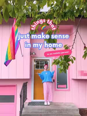 A photograph of a pink house with a person standing outside. There is a rainbow flag hanging on the left and text overlays over the image. The individual is smiling and looking at the camera with their arms open and wearing a blue sweater and striped pink pants.