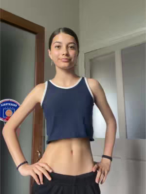 A medium shot features a young woman in dark, high-waisted athletic shorts and a navy crop top with white trim, posing with her hands on her hips. The background shows a partially open door and a glimpse of a room with a door and a basketball hoop. The overall composition is well-lit, with the focus on the model's mid-section.