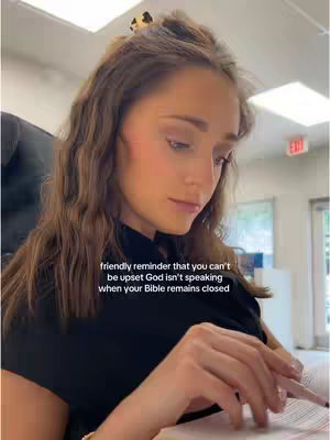 A woman with long brown hair is looking down at a Bible in her hands. She wears a black top and has a hair clip on her hair, the background is a well-lit room.