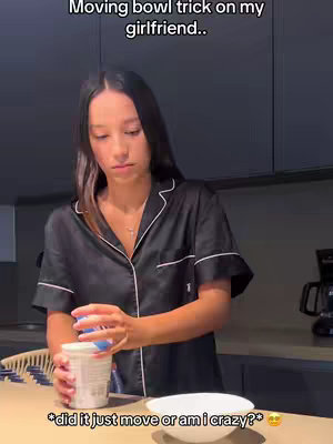 A woman in a dark silk pajama set is standing in a kitchen, looking down at a cup she is holding. She has long dark hair and a serious expression. In the foreground, a white bowl sits on a wooden countertop. The background is a modern kitchen with dark cabinetry and a countertop. Text overlays are present, providing context or commentary.
