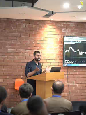 A man stands at a podium in front of an audience, presenting with the projected screen in the background. A laptop is on the podium, and an orange chair is visible. The background is a exposed brick wall.