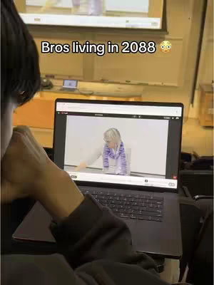 A person is shown from the shoulders up, with their arm and hand resting on the laptop, obscuring most of the rest of their body. The laptop screen displays a video of a person in a lecture hall. Above the laptop an eye level of the camera is a wider view of lecture hall, with a projector screen and a few other details. Text is overlaid on the image.