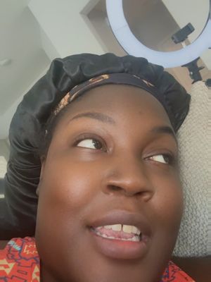 A close-up selfie of a person with dark skin wearing a black satin sleep cap and a colorful top. The person is looking up to the right, with eyes slightly widened and a slight smile. A ring light is visible in the background, and the setting appears to be indoors.