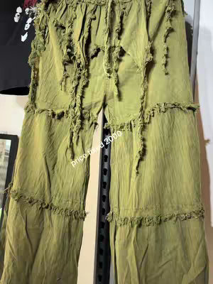 The image features a pair of olive-green pants hanging with a dark colored t-shirt in the background. The pants are the central focus, showcasing a unique design with frayed edges that give them visible texture and an aesthetic appearance. The pants are hanging against a metallic shelf, and the text 'price $2000' are written on the pants.