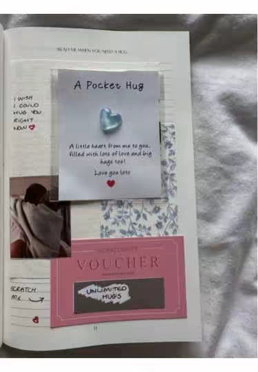 The image shows an open notebook page with a note and other elements attached. A blue heart-shaped object sits on top of a white card with text 'A Pocket Hug'. Below that is a pink voucher, and to the left, a photo and handwritten notes are visible.