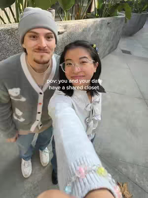 A selfie-style image featuring a couple posing closely. The woman in the foreground is holding the camera, while the man stands to her left. Both are smiling and appear to be in a casual setting, possibly outdoors. The background shows a concrete wall and some greenery.