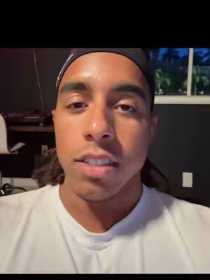 A selfie of a man in a white t-shirt and a black cap, facing the camera with a neutral expression. The background is dark, possibly a studio or room, with a window on the right showing some greenery. The lighting seems consistent, and the composition is simple and direct.