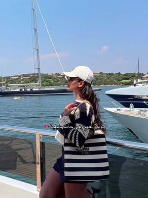 A woman wearing sunglasses and a white cap stands on a yacht deck, with a striped tote bag and a blue and white sweater. In the background, there is a body of water with boats and the sky is clear.