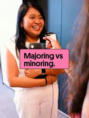 A woman with black hair and a white top is holding a microphone, smiling. The background includes a blue wall. A pink rectangle with text stating "Majoring vs minoring." is overlaid on the image.
