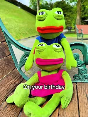 Two plush frog toys are sitting on a park bench. One is on top of the other, with the bottom one wearing a pink dress and the top one wearing a blue shirt. The background is a grassy hill with trees and a park bench visible.