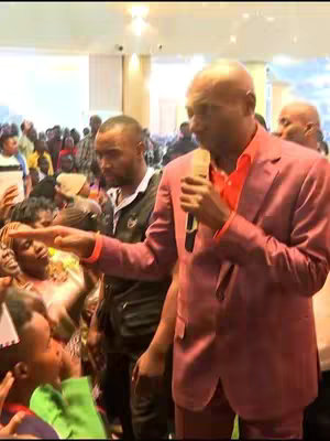 The image shows a man in a pink suit holding a microphone and speaking to a crowd, with another man standing beside him. The setting appears to be indoors, likely a church or auditorium. The man in the pink suit is gesturing towards the crowd with his hand.