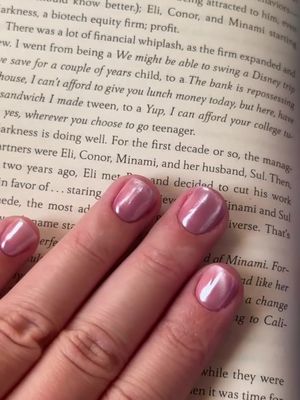 Close-up shot of a hand with short, polished pink nails against a backdrop of a book with text. The nails are the focal point.