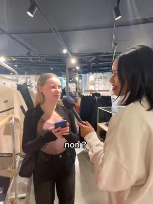 Two young women are positioned in a well-lit retail environment, possibly a clothing store. One woman, standing on the left, is holding a smartphone, and is being interviewed by the other woman on the right, who is holding a microphone. The woman on the left is the primary focus of the shot. The background is softly blurred, featuring clothing racks and store displays. The overall lighting is bright and neutral.