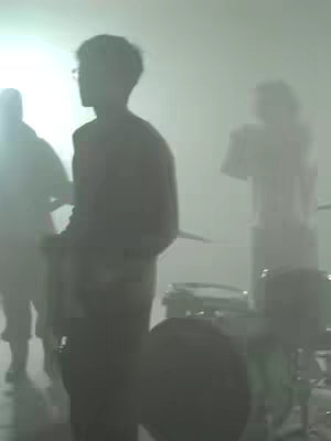 A blurry, low-contrast image of a band performing on a stage. The shot is dimly lit, with the band members appearing as silhouettes against a bright, diffused light source in the background. The focus is on the band, with a drummer and two guitarists are visible. The overall composition creates an atmospheric and mysterious feel.