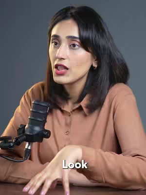 A woman with dark hair and a brown shirt sits in front of a dark background, speaking while holding a microphone. The word "Look" is overlaid on the image in white text.