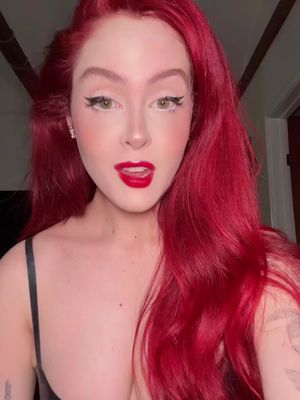 A close-up shot of a woman with bright red hair and bold makeup. Her hair is styled in voluminous waves that frame her face. She has fair skin, accentuated by eye makeup, including dramatic black eyeliner and white detailing, and a bright red lipstick. Her open mouth suggests she is speaking. She is wearing a black top.