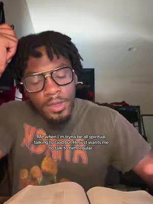 A man with glasses is positioned in front of a blurred background. He's in a close-up, with the focus on his face and expression. The man wears glasses with a black-framed frame and a dark-colored, short-sleeved shirt. His hair is styled in short dreadlocks. Below the man, there is an open book with white pages.
