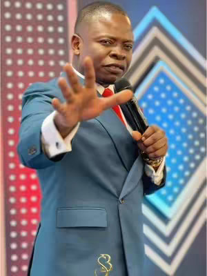 A man in a blue suit is holding a microphone and gesturing with his right hand, palm open. The background features a red, diamond-patterned stage design with some lights. The man is looking towards the camera, and the shot is well-lit.