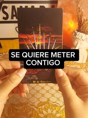 The image shows a person holding a tarot card with the text overlay "SE QUIERE METER CONTIGO". In the background, there's a blurred setting of a woman's hands holding the tarot card with a soft, warm light. There are also some other objects and setting to add an environment.
