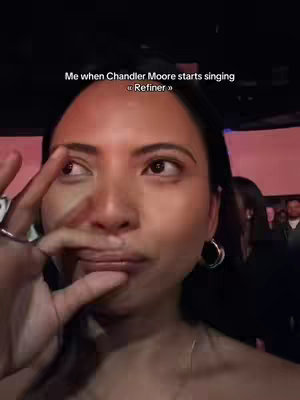 A woman is shown in a selfie-style shot, her face taking up most of the frame. She wears a black hoop earring in her left ear and touches her finger to her cheek, with some other people in the background, blurred out.