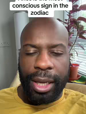 A close-up shot features a Black man with a bald head, closed eyes, and a dark beard. He wears a yellow shirt and is positioned against a softly blurred indoor background with a plant and window. Text above reads, 'Aries is the most conscious sign in the zodiac.'