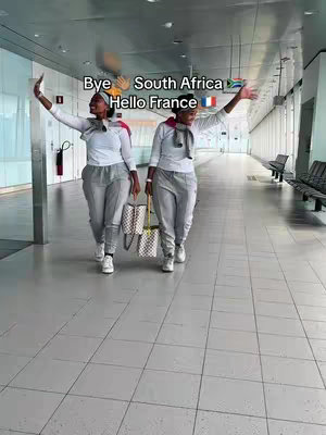 Two women, likely twins, are walking towards the camera in an airport corridor. They are waving with their arms up, one holding a patterned tote bag. Text overlays read 'Bye South Africa' and 'Hello France' with flag emojis, with a light gray background.