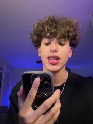 A young man with curly brown hair is holding a black phone in front of his face. He is looking up at the camera and his mouth is slightly open as if he is speaking. The background suggests he's in a room with purple and blue ambient lighting.