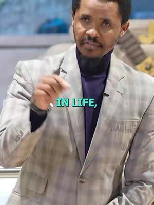 A medium shot of a man in a grey suit and purple turtleneck, gesturing with his right hand. Text overlay 'IN LIFE,'. The background appears blurred.