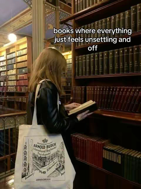 A person with long brown hair, wearing a black jacket and carrying a canvas tote bag, is standing in a library, surrounded by bookshelves filled with books. The person is holding a book open, and the text overlay indicates that it is a book recommendation.