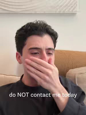 A young man covers his mouth with his left hand. He has short, dark hair and is wearing a black shirt. The background shows a light-colored wall and a beige couch. Text overlay at the bottom reads, 'do NOT contact me today.'