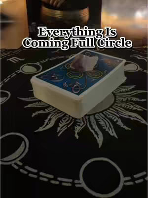 The image is a close-up shot of a tarot card deck resting on a patterned black surface. Overlaid on the deck is a small, clear crystal. The deck is the central focus, slightly angled to reveal its edges. In the background, partially visible, is a golden singing bowl above a black circular background with white symbols.
