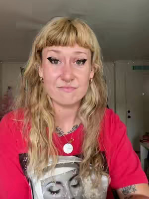 A woman with blonde hair and heavy dark eyeliner looks directly at the camera. She wears a red t-shirt with an image on it and a nose ring with a necklace.