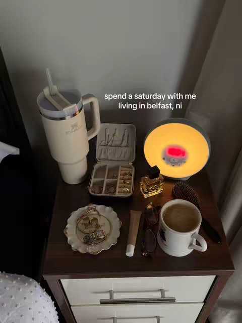 A well-lit shot of a bedside table with various items on top. A cream-colored mug filled with coffee, a white tumbler, an open jewelry case and a ring tray, perfume, sunglasses, and a wake-up light. The background is blurred, but appears to be a light-colored wall.