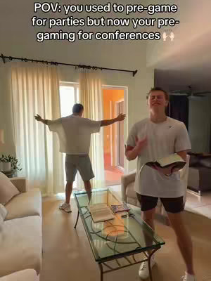 A room interior with two young men. One stands in the foreground holding an open book with a soft smile. The second man stands in the background with his arms spread open. The room has natural lighting. The background is a beige and white color, creating a cozy mood.