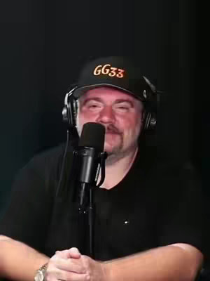 A man is centered in the frame, viewed from the chest up against an entirely black backdrop. He's wearing a black baseball cap, which has the orange text "GG33." He has a neatly trimmed mustache and goatee. He wears black headphones and a black shirt. A silver microphone is directly in front of him. His wrist watch and a portion of his hand are visible below the microphone. The image is well lit, putting the focus onto the man.