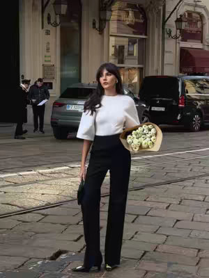 A woman in a white top and black pants stands on a cobblestone street, holding a bouquet of white flowers. Cars and buildings are in the background, indicating an urban setting.