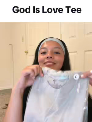 A woman is holding up a shirt in a plastic bag. The text "God Is Love Tee" is over the image. She is smiling at the camera.