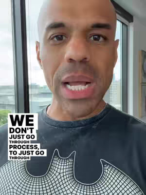 A bald man with dark skin is centered in the frame, speaking to the camera with his mouth open. He wears a dark gray t-shirt featuring a white graphic that appears to be the iconic Batman logo. Text overlays are positioned in the lower left corner. The background shows a window with an outdoor view of a cityscape and sky, suggesting an indoor setting with natural light.